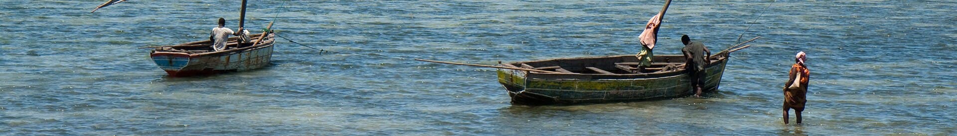 Two boats with rolled-up sails in Mozambique. Both has a man in them, and another is standing in the sea next to one boat.