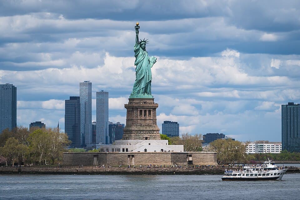 The Statue of Liberty (Bartholdi, 1886) on Liberty Island; a sightseeing boat passes in the foreground with the New Jersey skyline behind.